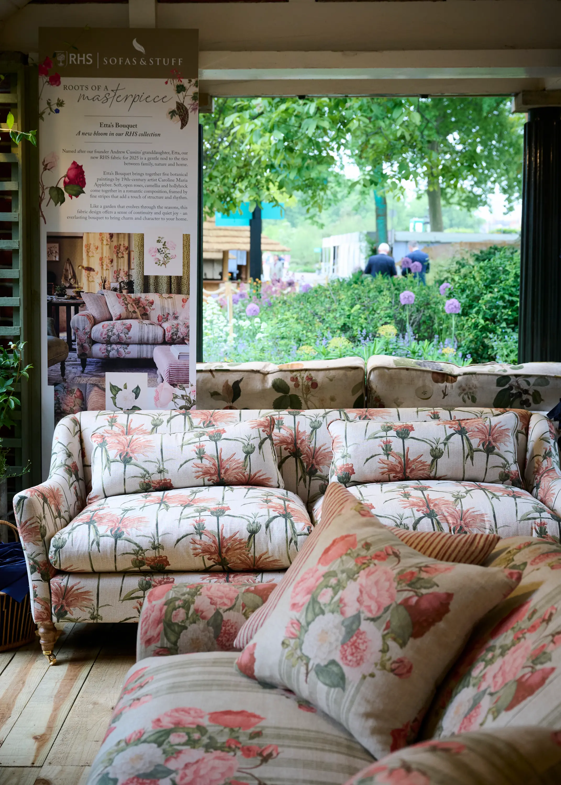 Floral-upholstered sofas and cushions displayed inside the RHS Sofas & Stuff stand at the Chelsea Flower Show, with garden planting visible through the open window behind.